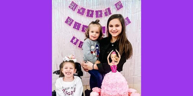 Three young sisters are sitting near a birthday cake.