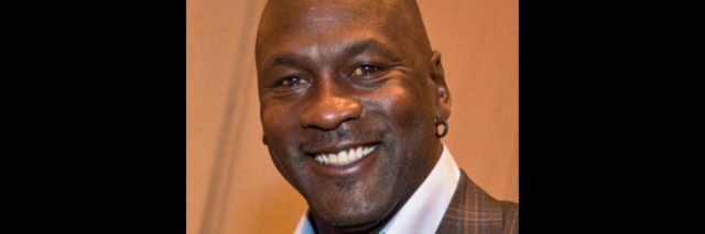 Michael Jordan, in a plaid brown suit, smiles at the camera