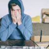 photo of man in hoodie sitting on sofa with his hands over his eyes, looking overwhelmed