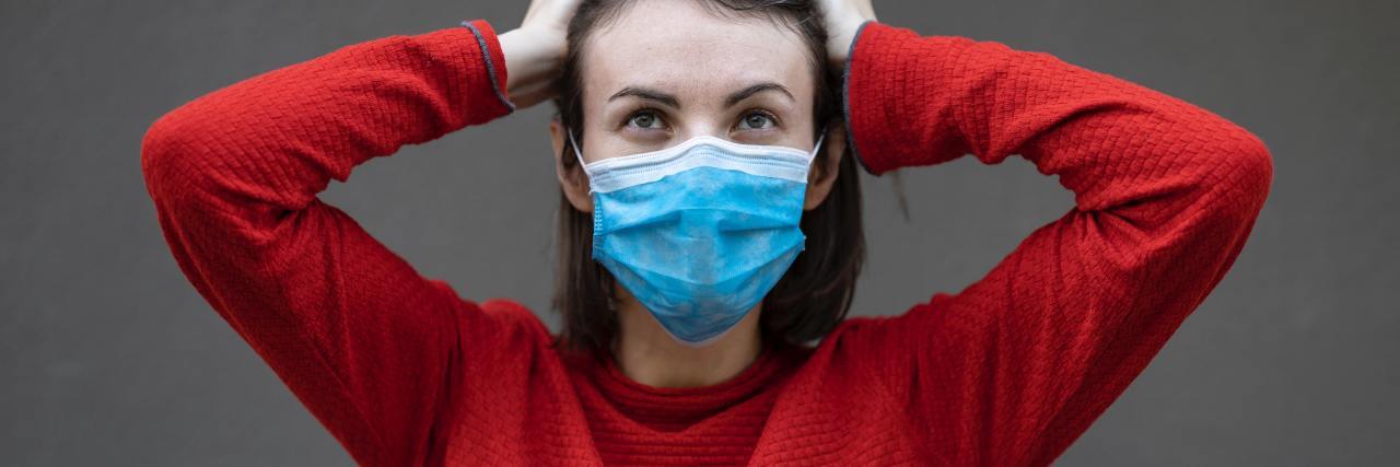photo of woman in red sweater, wearing blue medical mask, with hands raised to her head