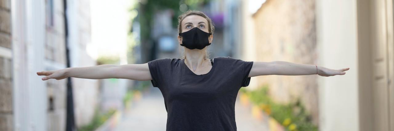 photo of woman standing in street with arms outstretched while wearing black face mask