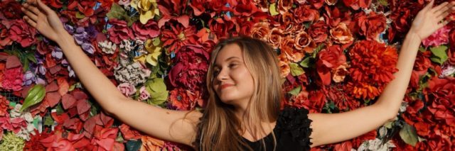 Woman with arms open wide in front of a wall of flowers