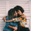 Two black women with natural, curly hair, one with her arms around the other