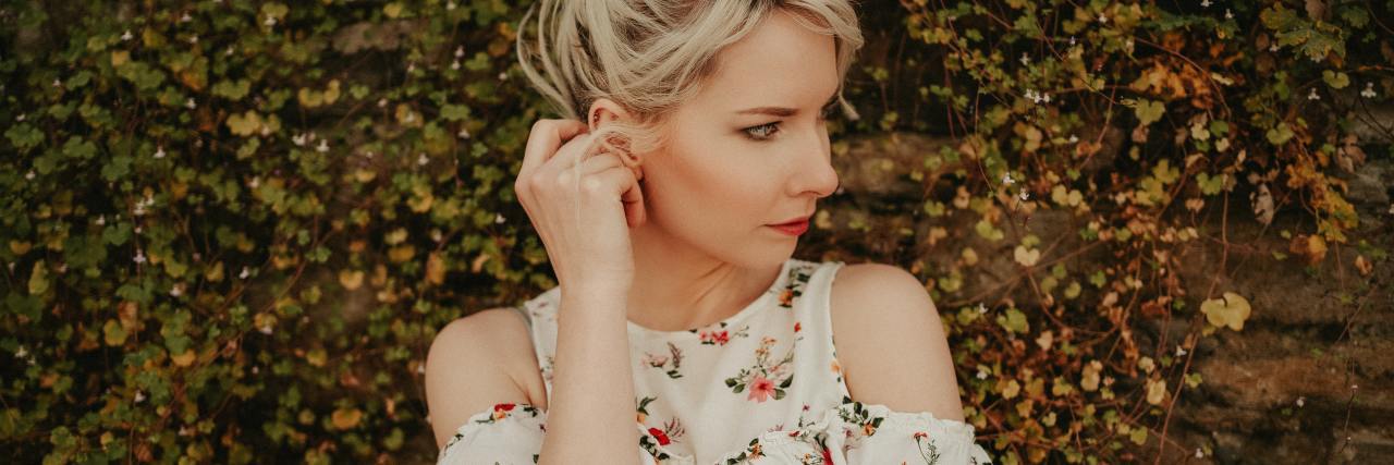 My Kid's Reaction to a Doctor's Appointment During COVID-19 photo of worried looking woman standing against leafy hedge background, one hand raised to the back of her head