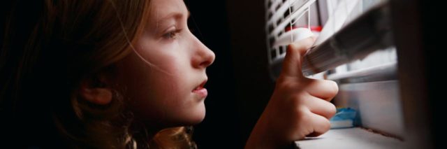 close up photo of girl peeking out of window blinds while inside is in darkness