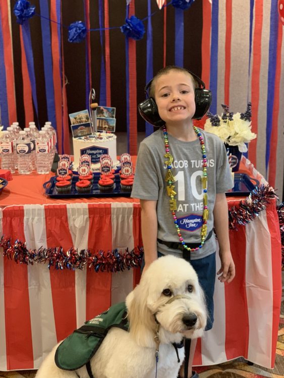 A boy with autism is smiling at his birthday party standing by a table with a cake and cupcakes.