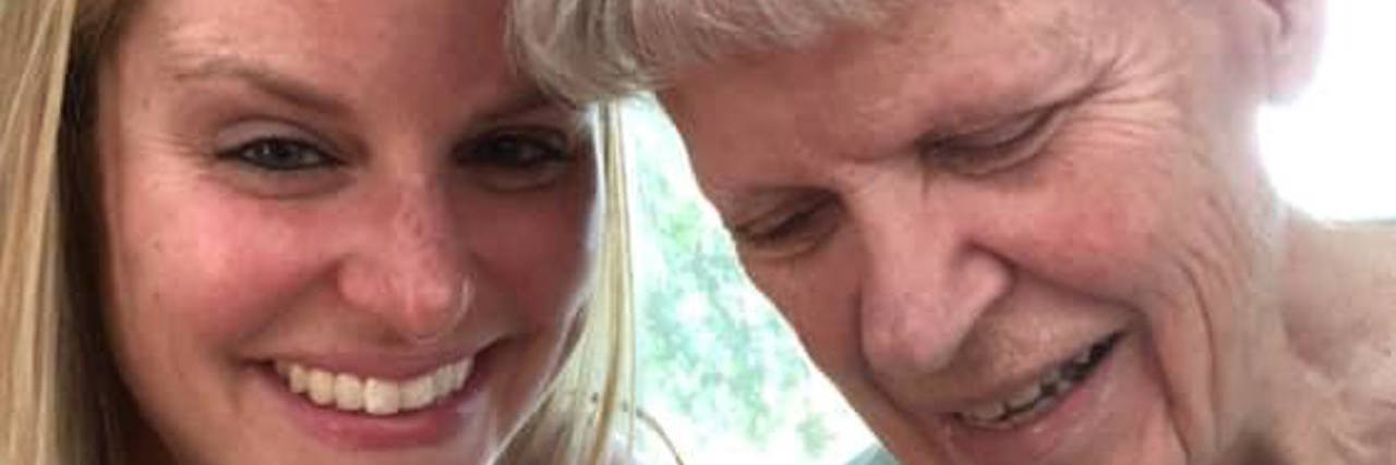 Caring for a Parent With Alzheimer's Disease: What It's Like Smiling younger woman sitting beside an older woman with short white hair who is looking down, both close together in a bright indoor setting.