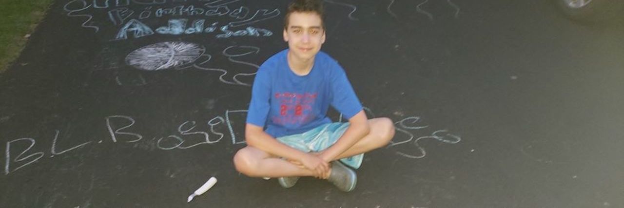 Autistic 'Birthday Lawn Bandit' Surprises Kids in Quarantine Birthday Lawn Bandit, a young white autistic boy sitting cross-legged on a driveway