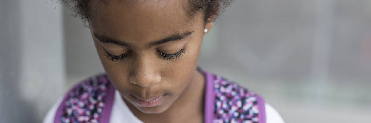 A young black girl looking down, wearing a backpack