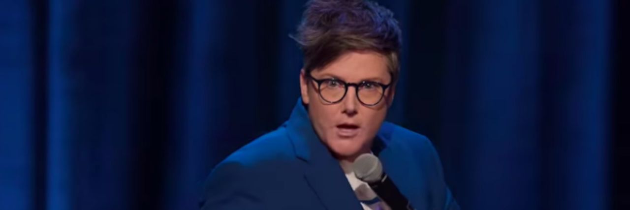 Hannah Gadsby, a white woman with short brown hair, glasses and wearing a blue blazer, speaks into a microphone