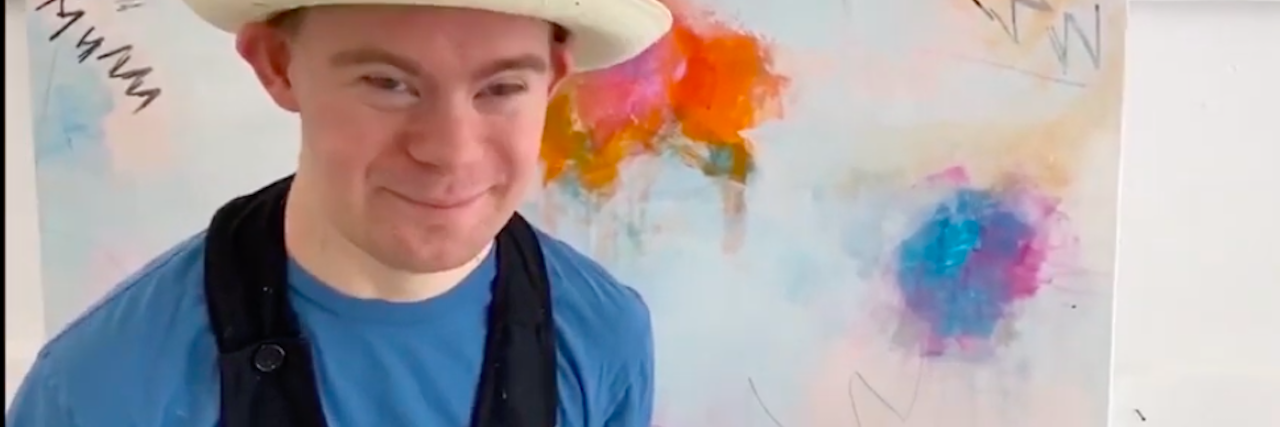 Artist with Down syndrome wearing a hat and standing in front of a canvas with paint on it