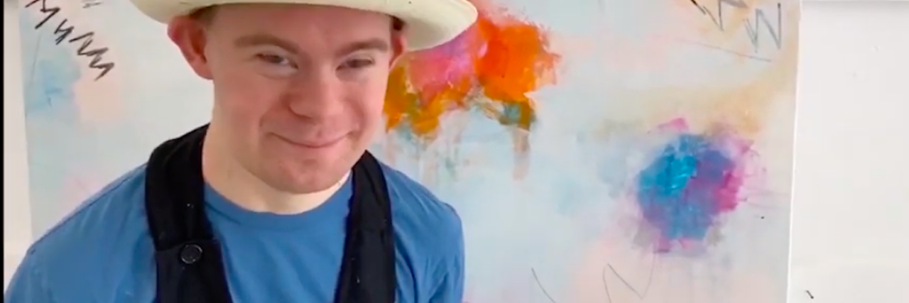 'Artists in Lockdown' Shows Artists With Down Syndrome During COVID-19 Artist with Down syndrome wearing a hat and standing in front of a canvas with paint on it