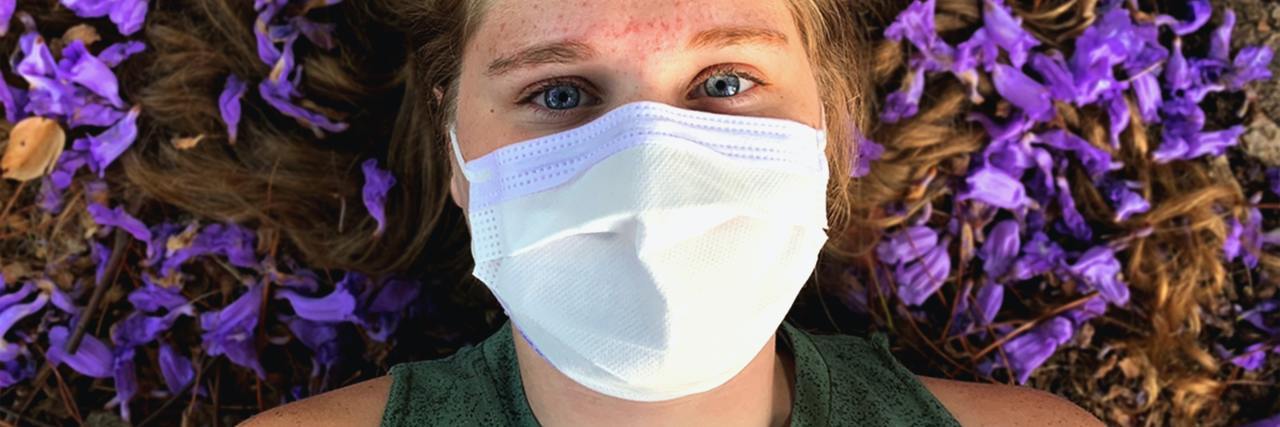 photo of woman lying on purple flowers, wearing a face mask and looking up into the camera