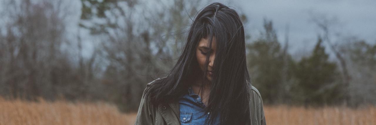 Study Suggests Fibromyalgia Patients Have Greater Risk of Self-Harm Young woman outside in a field wearing a denim shirt and jacket looking at the ground with her face partially covered by long brown hair