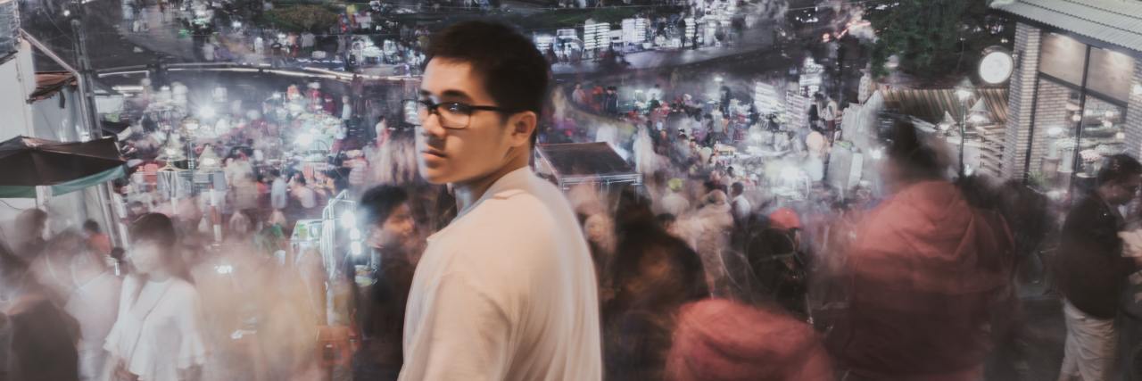 photo of young asian man standing in front of blurred people and looking into camera