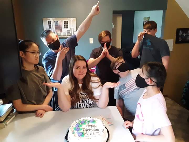 A group of people gathered around a birthday cake in a kitchen, wearing masks