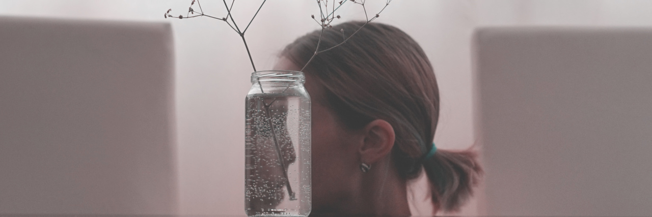 photo of woman whose face is partially hidden and reflected in a jar with a sparse plant