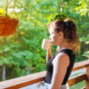 Woman standing on porch of house drinking tea or coffee.