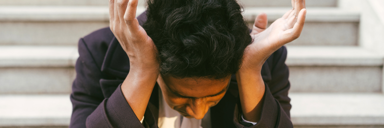 Being in Higher Education and Experiencing Stress and Mental Illness Young man with black curly hair sitting on steps with his hands to his ears, looking down. wearing a black jacket and white shirt.
