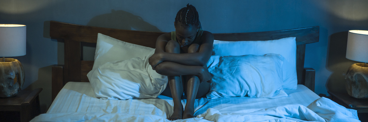 Young woman with her arms wrapped around her knees in bed in a partially lit room