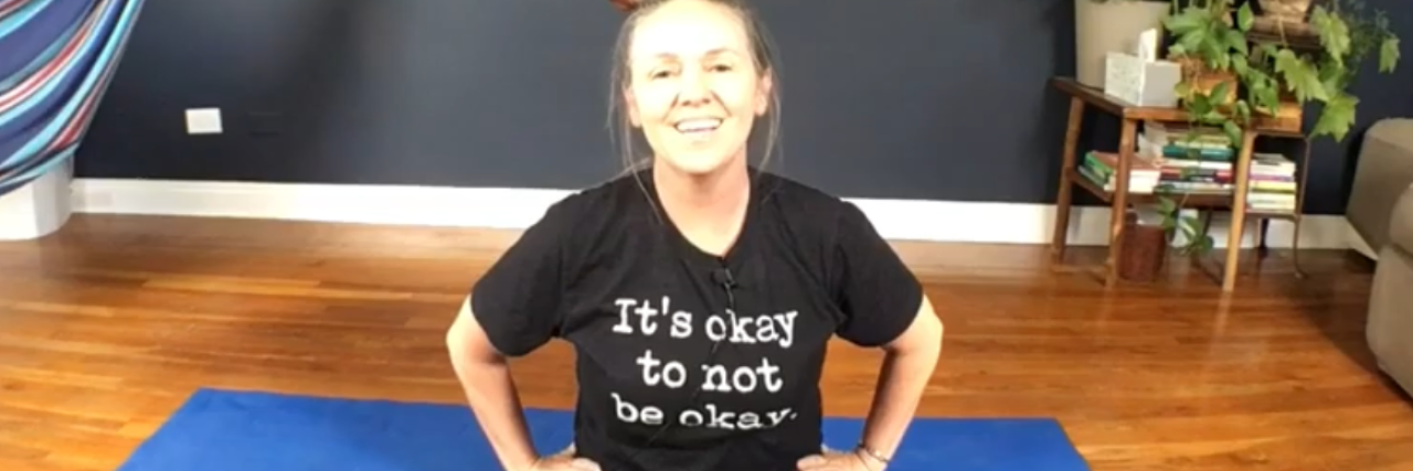 Yoga and Meditation Practice for Anxiety During the COVID-19 Pandemic White woman sitting on the floor smiling in a t-shirt that says "It's okay to no be okay."