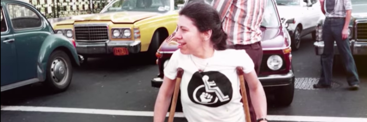 ‘Crip Camp’ Filmmakers Offer Free Panel Discussion Woman wearing a white shirt with a wheelchair on it using crutches