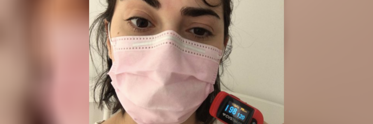 A woman wearing a pink mask, with a Pulse Oximeter on her finger