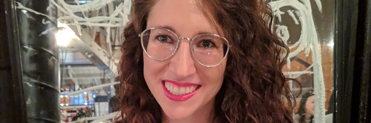 7 Myths About Blindness Jennifer at a restaurant. She has brown curly hair and glasses, and there's a panel of etched glass behind her.
