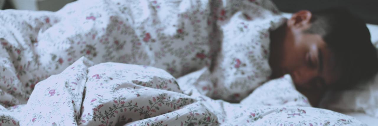 Why You Need to Prioritize Sleep During the COVID-19 Pandemic A man asleep under a comforter with his dog's head peeking out from under the blanket next time him