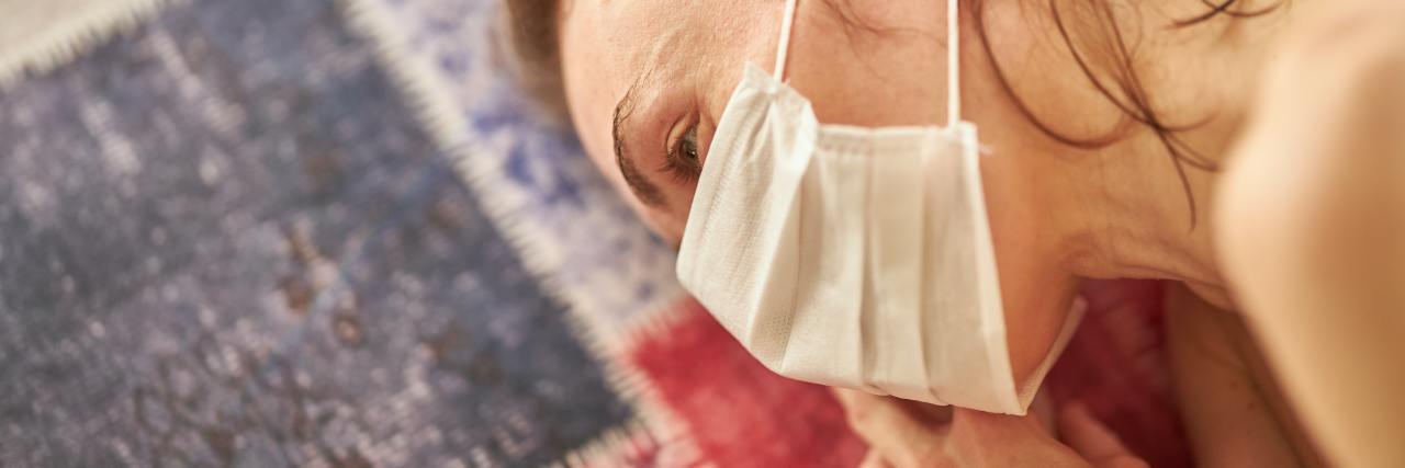 COVID-19 Quarantine's Affect on My Chronic Pain From Fibromyalgia photo of blonde woman lying on floor, wearing a coronavirus mask and staring into space