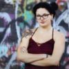 photo of woman with dark hair and glasses standing against graffiti wall with arms crossed, looking into camera