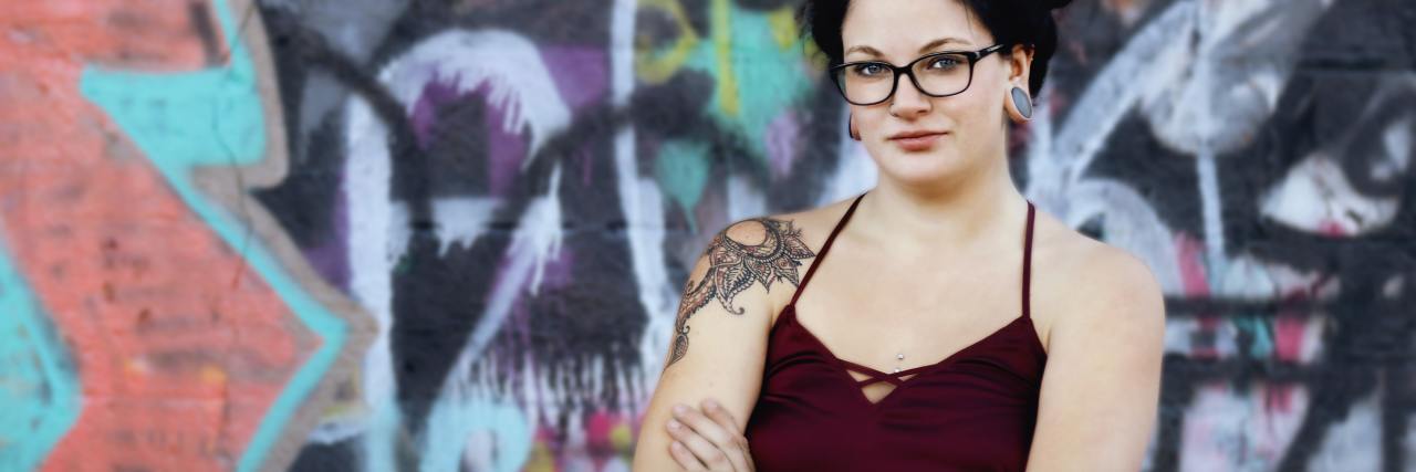 photo of woman with dark hair and glasses standing against graffiti wall with arms crossed, looking into camera