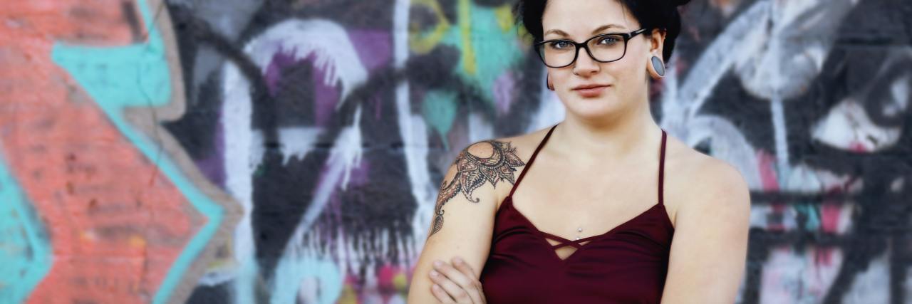 Feeling Ready To Face My OCD for the First Time photo of woman with dark hair and glasses standing against graffiti wall with arms crossed, looking into camera
