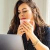 photo of woman in front of laptop with a mug raised to her face