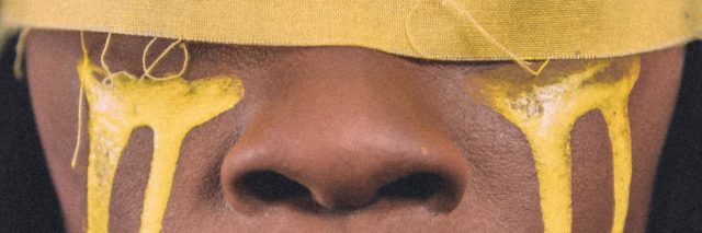 photo of Black woman with yellow covering over her eyes and yellow paint running down her cheeks like tears