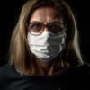 photo of older woman with glasses and blonde hair, standing in darkness and wearing a white face mask to protect against coronavirus