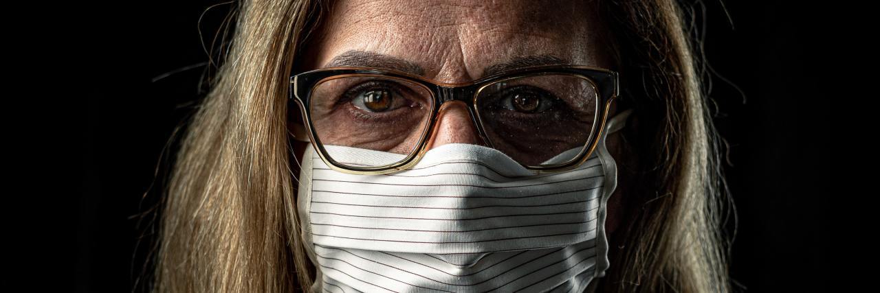 photo of older woman with glasses and blonde hair, standing in darkness and wearing a white face mask to protect against coronavirus