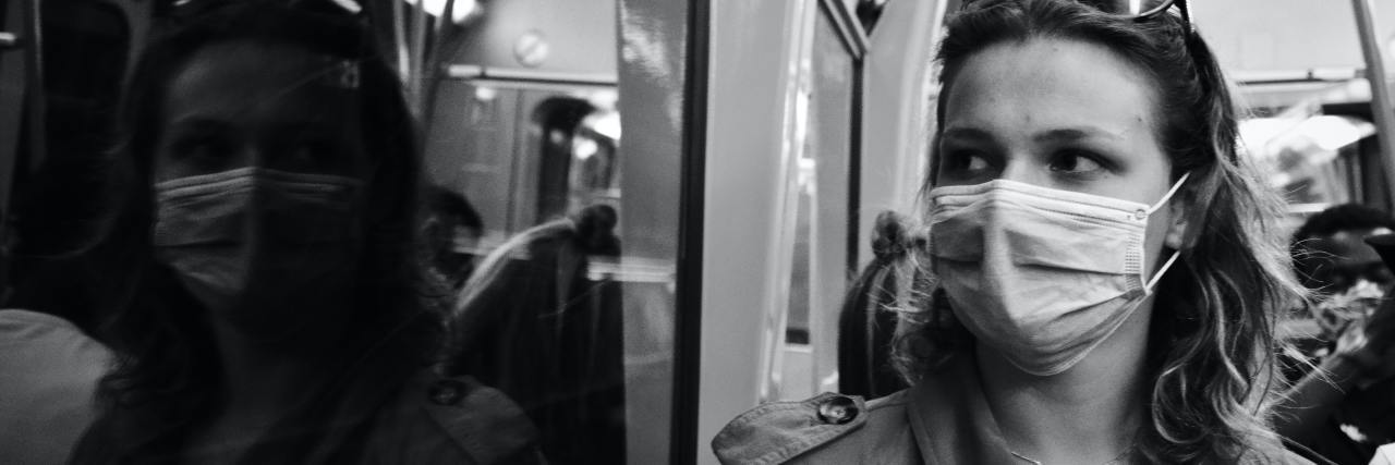 We Need to Feel Safe to Recover After COVID-19 Pandemic Trauma black and white photo of woman wearing a face mask looking out of train window with her reflection visible