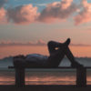 A man is lying on a wooden bench, a sunrise in the background