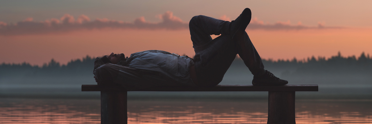 Life With Illness: Things That Seem 'Lazy,' but Aren't A man is lying on a wooden bench, a sunrise in the background