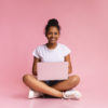 Student sitting cross-legged with an open laptop in her lap against a pink background