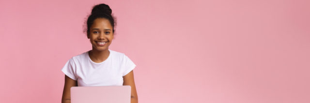 Student sitting cross-legged with an open laptop in her lap against a pink background