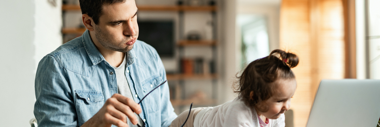 A dad on his computer trying to work. He has a stressed out look on his face as his child blocks the computer