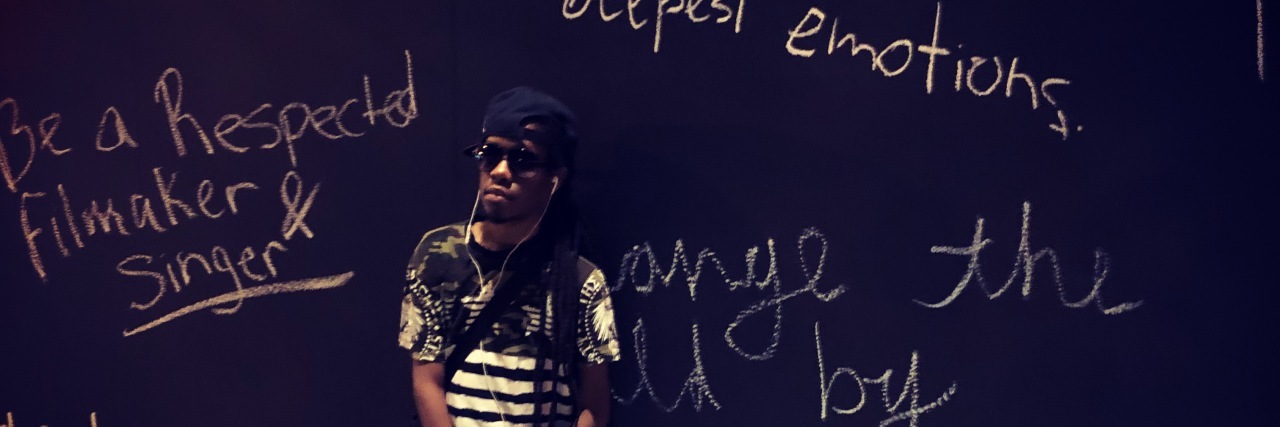 Kadeem standing in a black shirt against a chalkboard wall, wearing headphones and sunglasses