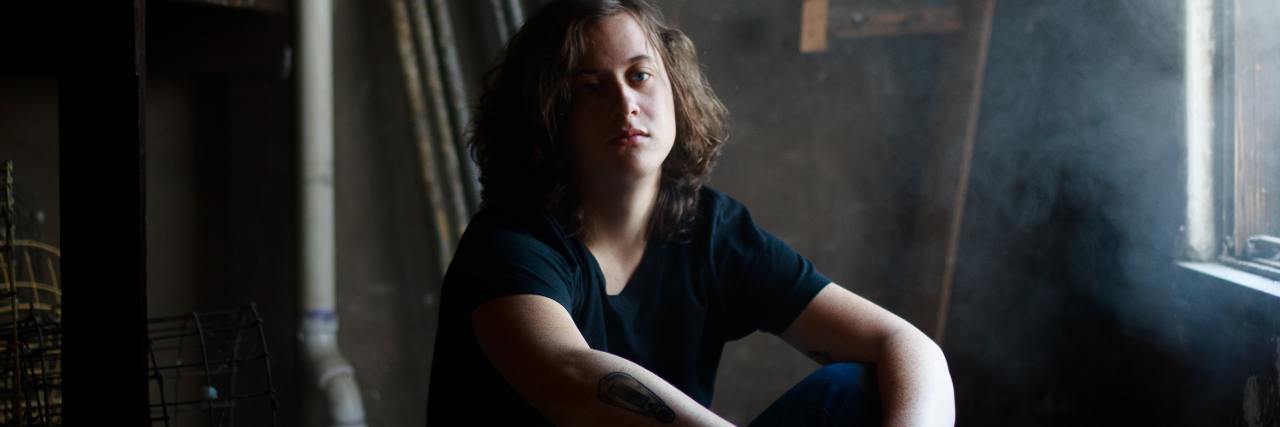Essential Depression Songs From the Past 25 Years photo of young man sitting in empty room with misty light coming through a window, looking into the camera, while he sits on the floor