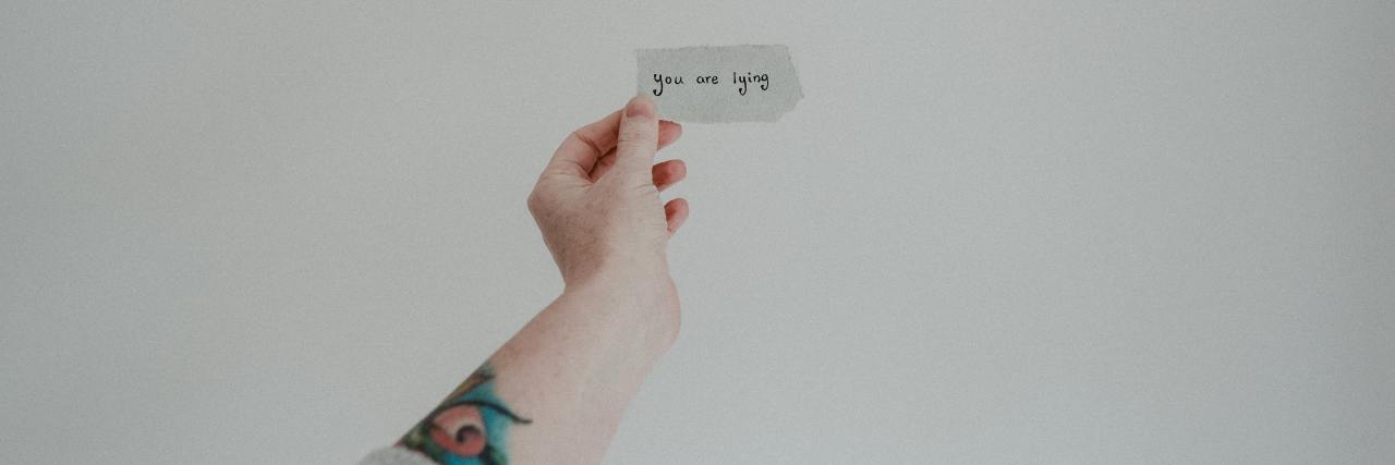 photo of an outstretched arm and hand, holding a note with reads "you are lying"