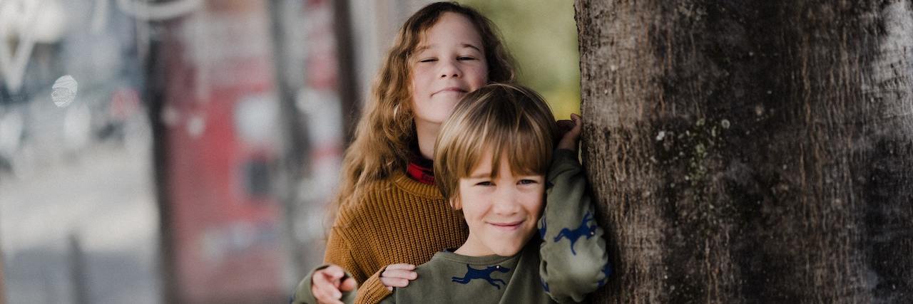 15 Childhood Experiences You Might Not Realize Affect You as an Adult Young girl and her younger brother leaning against a tree trunk