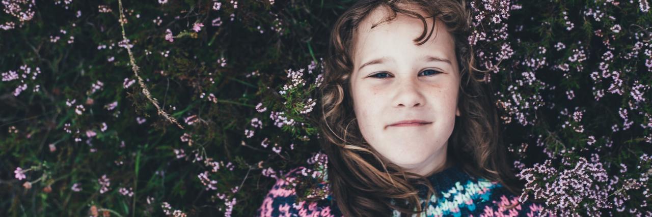 Can I Have Childhood Trauma If I Had a Happy Childhood? overhead photo of young girl lying on ground with flowers, looking into camera and smiling