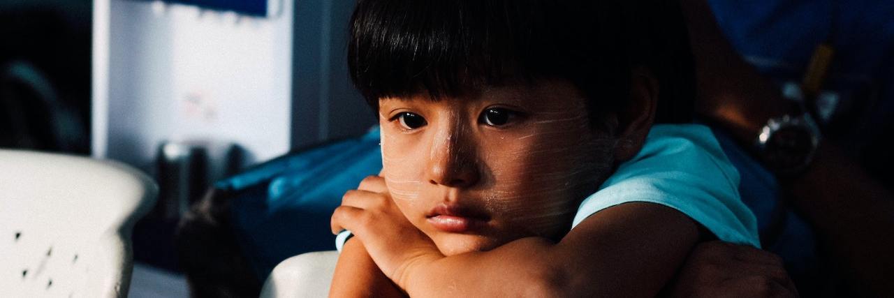 Study Suggests People Who Experienced Childhood Trauma May Age Faster Sad boy with dark brown hair with his head resting on his crossed arms leaning on a piece of furniture