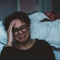 photo of woman sitting beside a bed, crying with her head resting on her hand